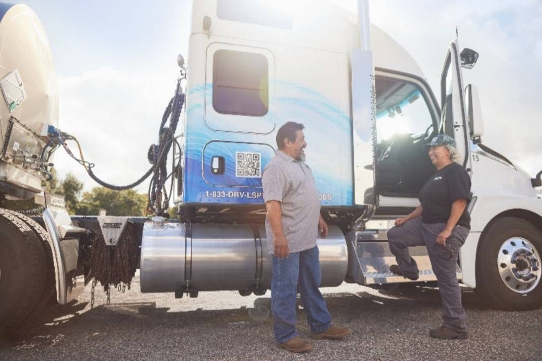 Photo of two drivers standing next to a semi-truck. One driver is male and one driver is female. They are having a conversation and are smiling.