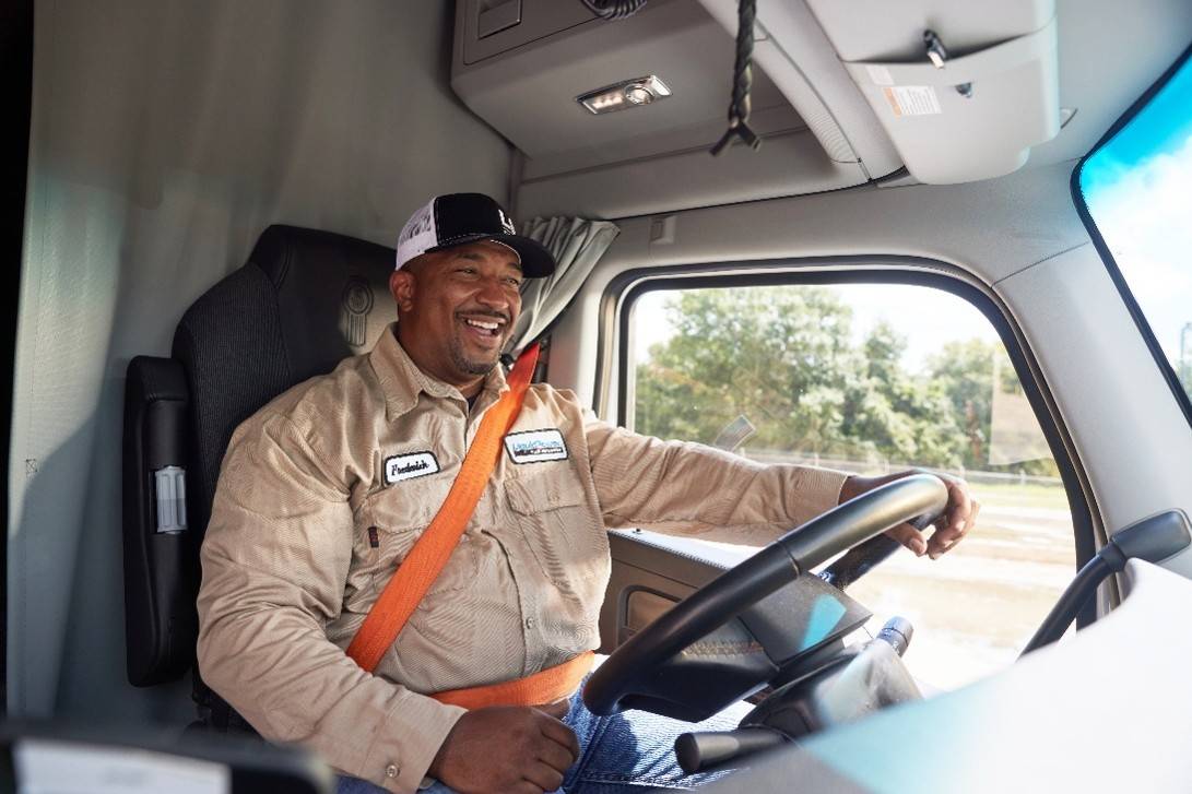 A male professional commercial semi-truck driver is actively driving a semi-truck. He is smiling. He is wearing an orange seat belt for safety.